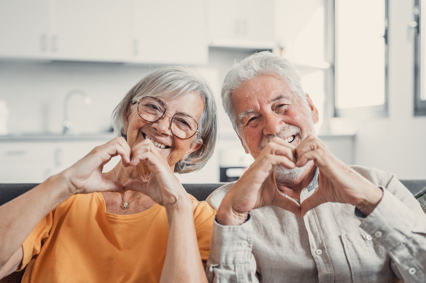 Bild eines Rentnerpaares, das mit den Händen das Herzzeichen formt und in die Kamera lacht.